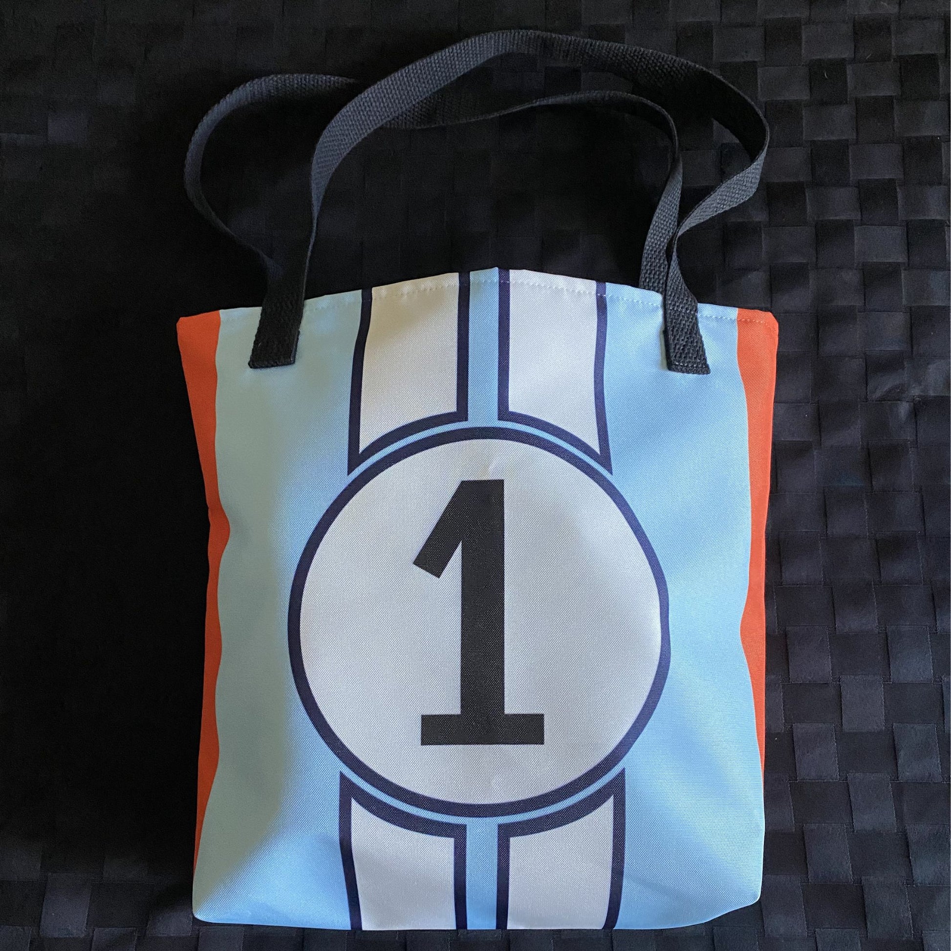 Blue tote bag with a number 1, racing stripes and orange accents on a textured black surface