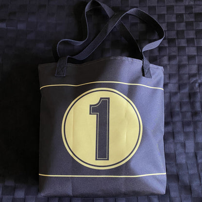Black tote bag with gold stripes, a gold circle and number one on a textured black surface