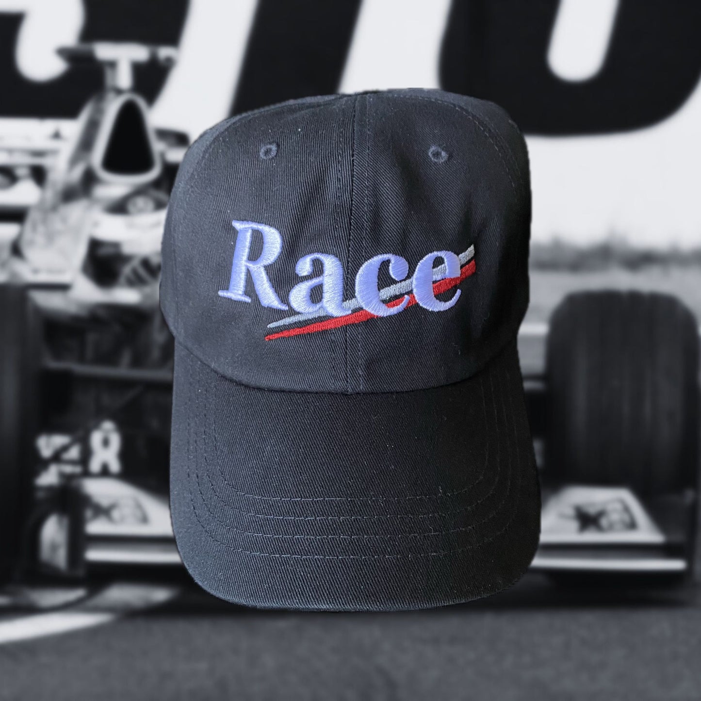 Black racing hat with 'Race' text in white with silver and red stripes on a blurred race car background