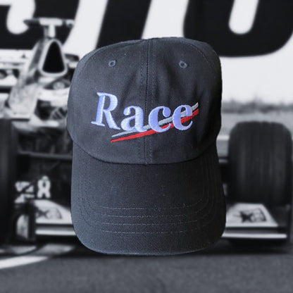 Black racing hat with 'Race' text in white with silver and red stripes on a blurred race car background
