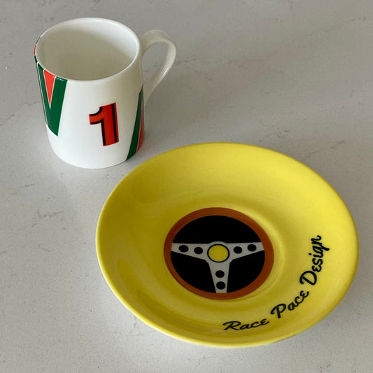 Yellow saucer with steering wheel design next to a race car coffee cup with red and green accents on a gray surface.