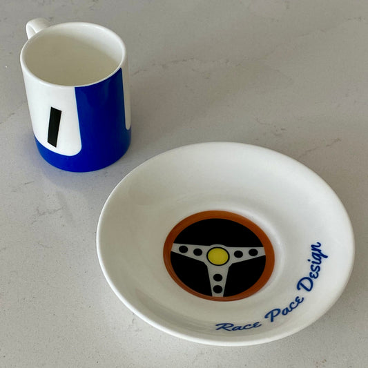 White cup with blue accents and a white dish with a steering wheel design on a light gray surface