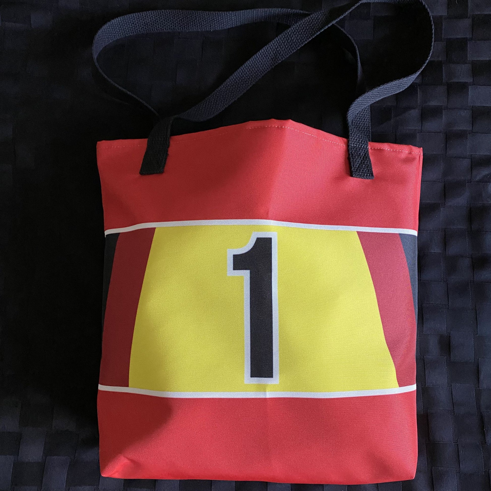 Red tote bag with a black, red and yellow stripe with a black number 1 on a textured black background