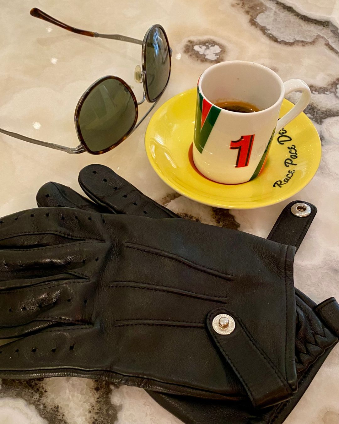 Automotive racing-inspired espresso coffee cup and saucer laid on marble surface next to driving gloves and sunglasses
