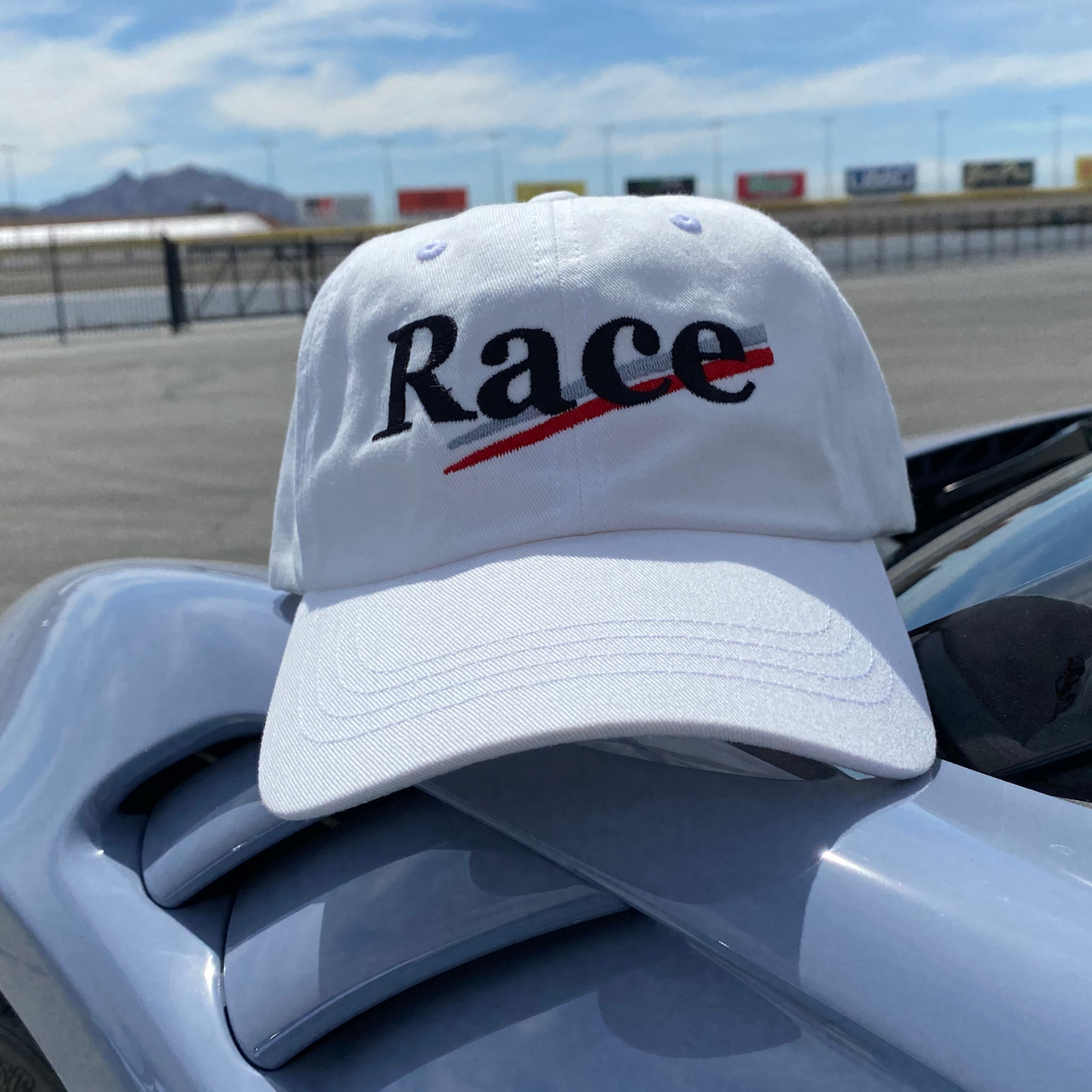 White cap with 'Race' text in black with silver and red stripes on a car at a race track